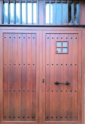 Puertas dobles de madera con tachuelas decorativas y una pequeña ventana, rematada por una ventana enrejada.
