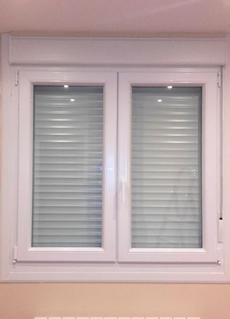 Ventana blanca de doble panel con persianas gris claro cerradas y un marco blanco, frente a una pared blanca y beige.