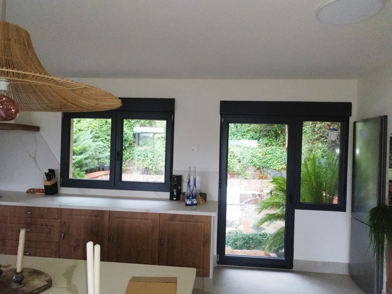 Cocina con ventanas y puerta con marco negro que dan a un exuberante patio trasero. También se ven gabinetes de madera y una lámpara de tejido.
