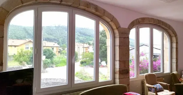 Dos ventanas arqueadas con marcos blancos y marcos de piedra ofrecen vistas a un pueblo y a las montañas. El interior cuenta con paredes y asientos beige.