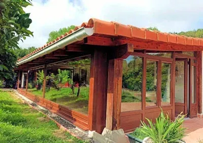 Terraza acristalada al aire libre con estructura de madera, techo de tejas rojas y paredes de cristal, con vistas a un césped verde.