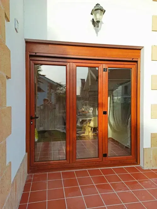 Puertas de patio de vidrio con marco de madera y suelo de baldosas. Las puertas reflejan el patio exterior. Una lámpara decorativa se encuentra encima.