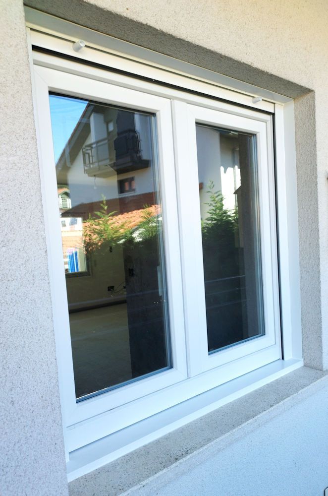 Ventana blanca de doble acristalamiento con superficie reflectante, ubicada en la pared gris claro de un edificio. El vidrio refleja partes de una casa y árboles.