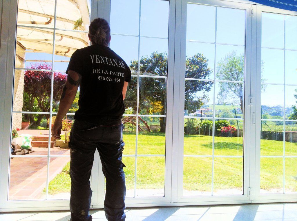 Persona con camisa negra y jeans parada frente a grandes ventanales con vista a un patio soleado.