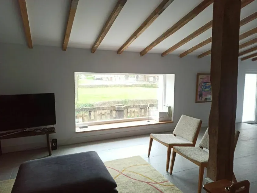 Sala de estar con gran ventanal, vigas de madera y muebles minimalistas. Tonos neutros con vistas a la zona verde.