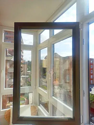 Ventana abierta con marco marrón que revela una vista exterior de los edificios y el cielo. Marco interior blanco visible.