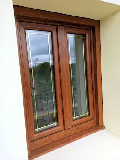 Una ventana de madera de dos paneles con una reja decorativa amarilla. Está enmarcada por un marco marrón y se encuentra en una pared blanca.