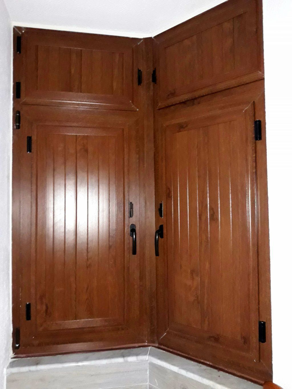 Persianas de madera marrón cerradas en una ventana esquinera. Las persianas tienen líneas verticales y bisagras y tiradores negros.