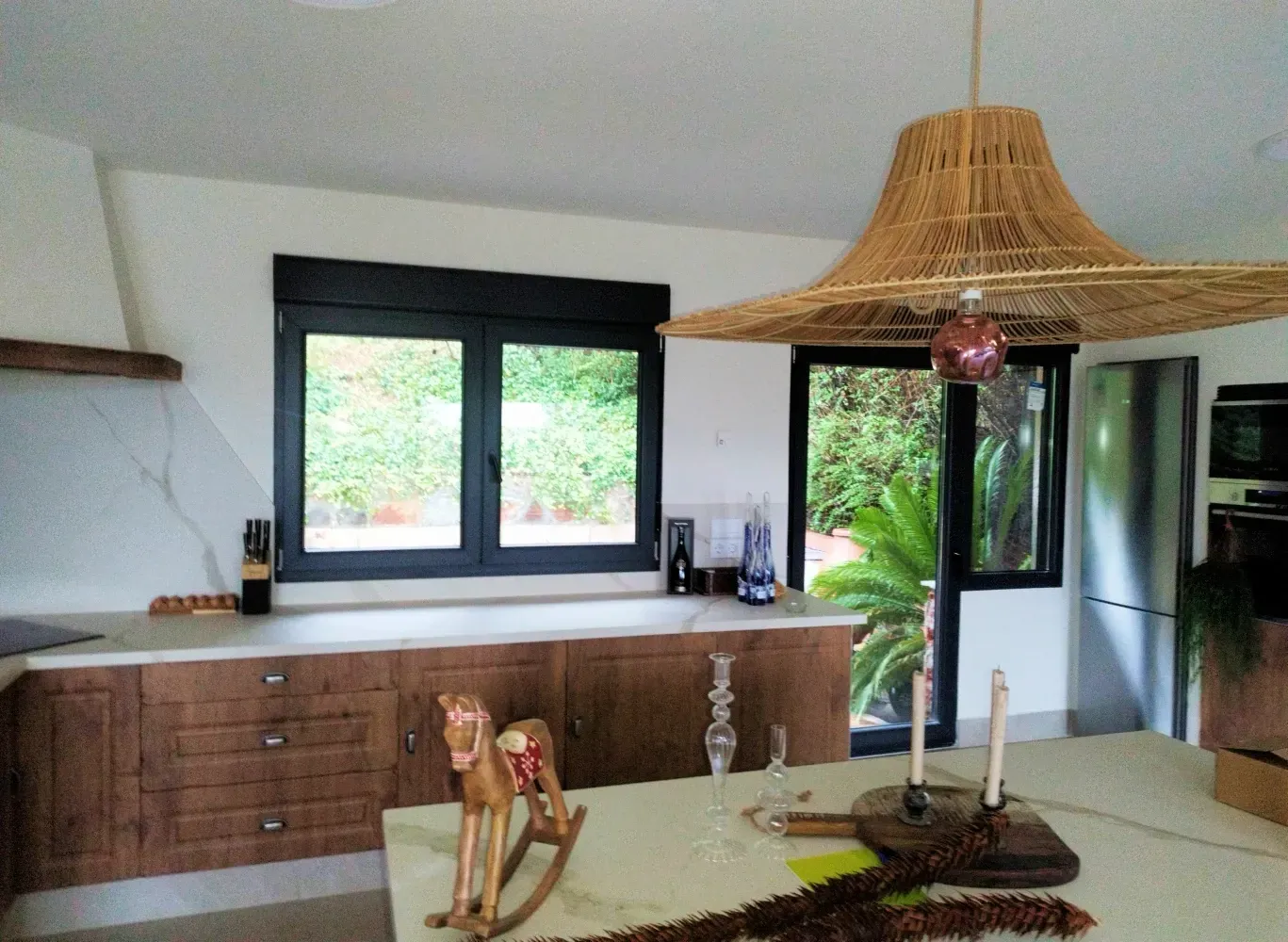 Cocina con gabinetes marrones, encimeras blancas y una gran pantalla tejida sobre el comedor. La ventana da a una exuberante vegetación.