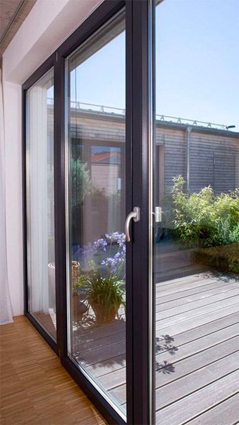 Una puerta corrediza de vidrio da a una terraza de madera con macetas y plantas. Un día soleado y luminoso.