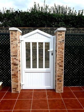 Puerta de jardín blanca con paneles de vidrio y techo decorativo, entre pilares de ladrillo, frente a una cerca de celosía y un seto verde.