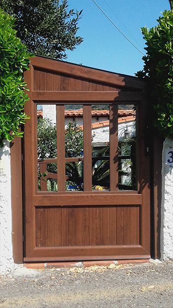 Puerta de madera marrón con cristales, colocada en una pared de piedra blanca, rodeada de arbustos verdes.