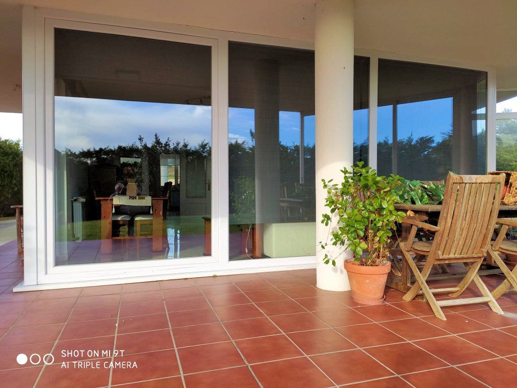 Vista de un porche con puertas corredizas de vidrio que reflejan el exterior. A la derecha hay una mesa y sillas, y junto a una columna blanca hay una maceta con una planta.