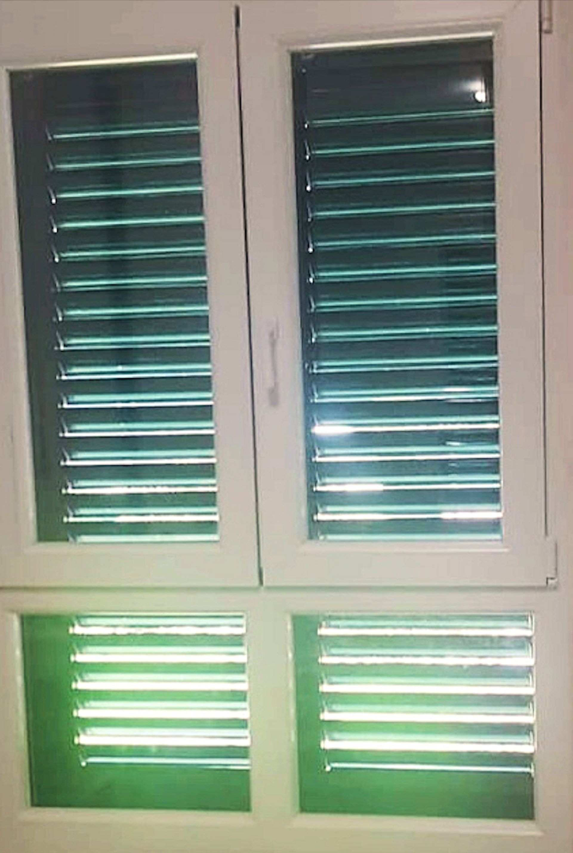 Ventana blanca con tres secciones de persianas horizontales teñidas de verde, probablemente en el interior. Dos secciones superiores y una inferior.