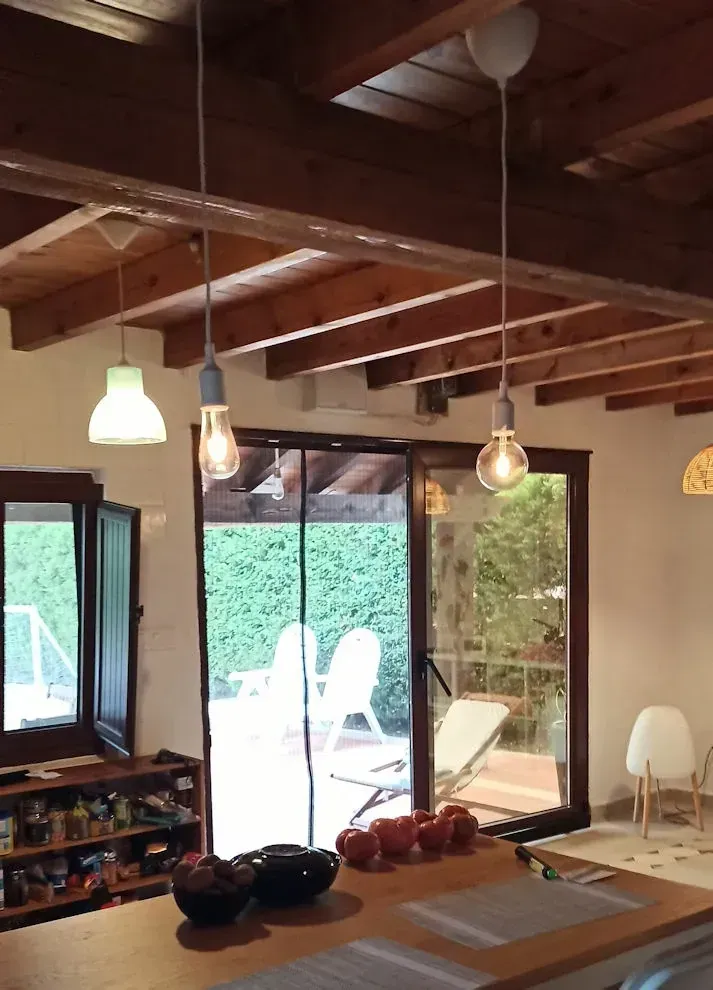 Interior de cocina con vigas de madera en el techo, luces colgantes y vista a un patio a través de puertas de vidrio.