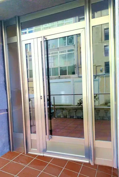 Una moderna puerta de entrada de vidrio con marco metálico. La puerta está abierta y refleja un edificio al otro lado de un patio.