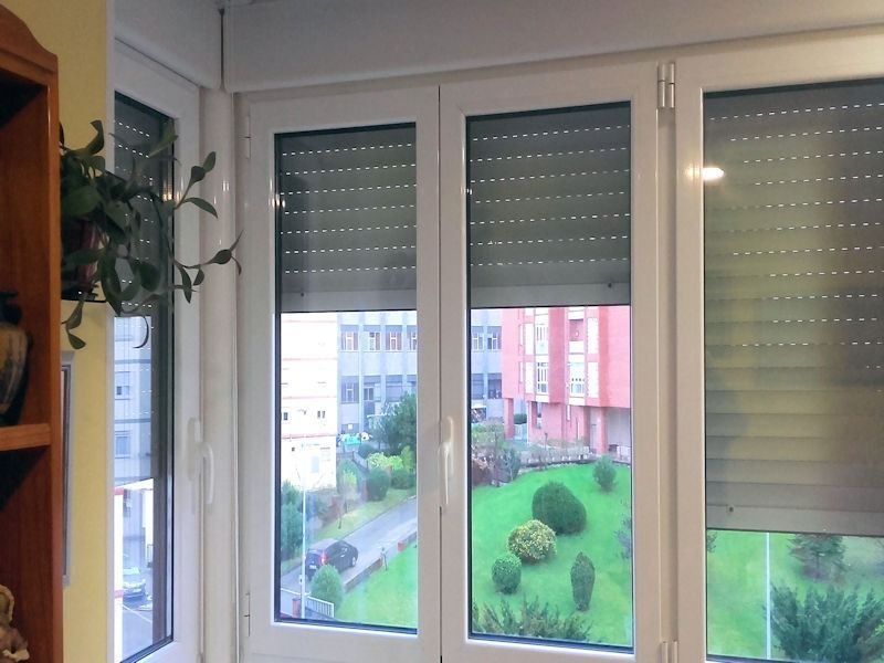 Una serie de ventanas con marcos blancos y persianas grises cerradas, con vista a un patio verde con edificios.