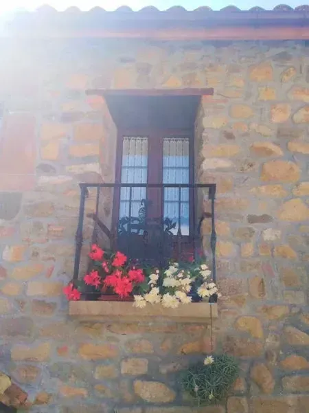 Balcón con flores rojas y blancas en un edificio de piedra. Barandilla negra y puertas dobles.