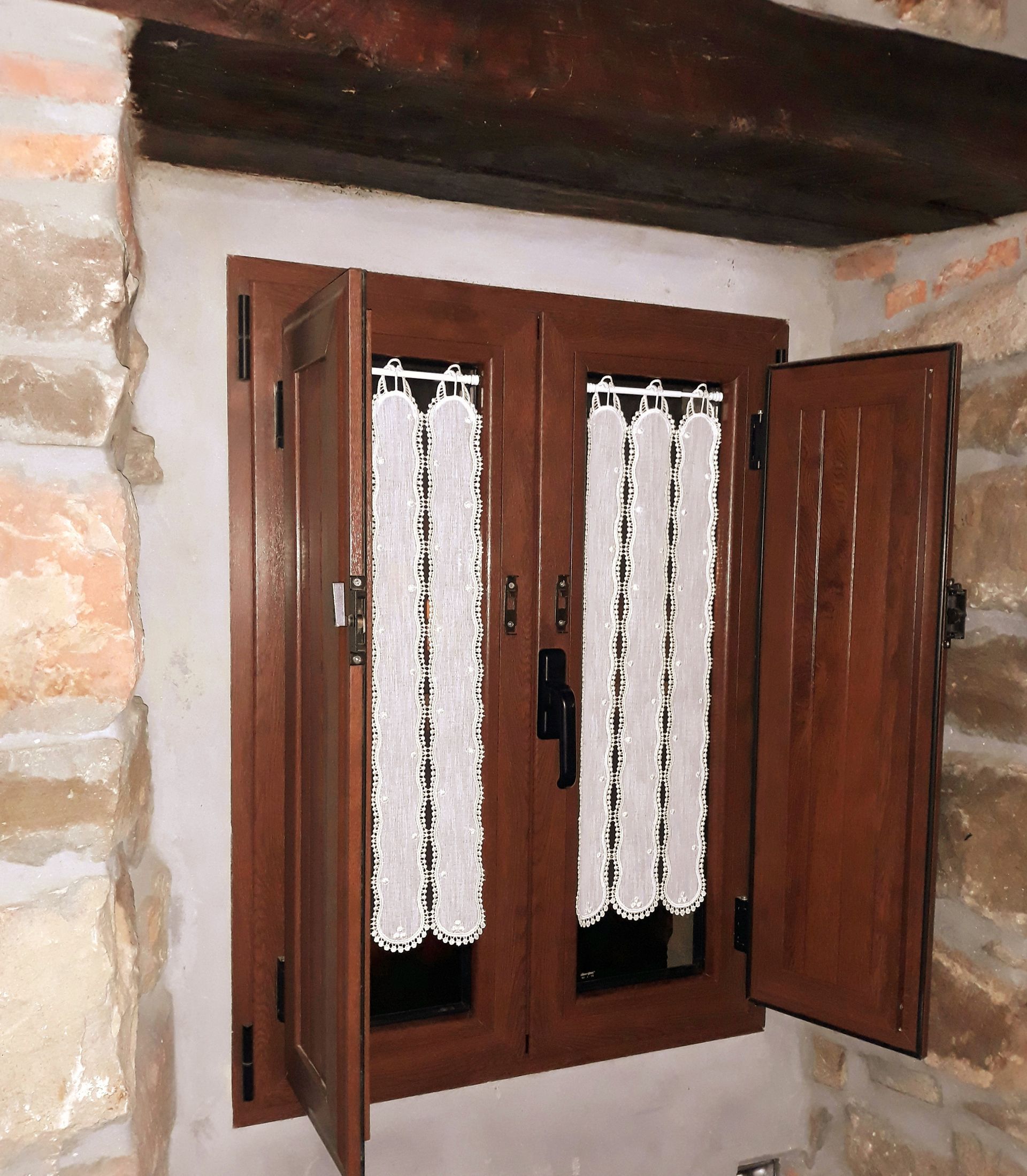 Ventana de madera con contraventanas abiertas y cortinas de encaje blanco, situada en una pared de piedra.