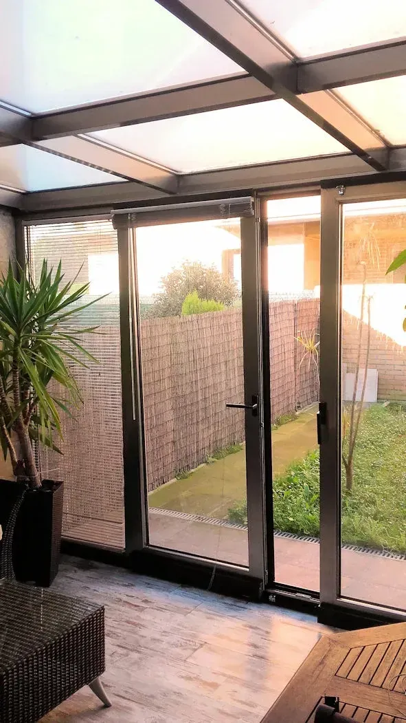 Terraza acristalada con paredes de cristal y techo transparente. A través del cristal se puede ver un patio trasero con plantas y una pared de ladrillo.