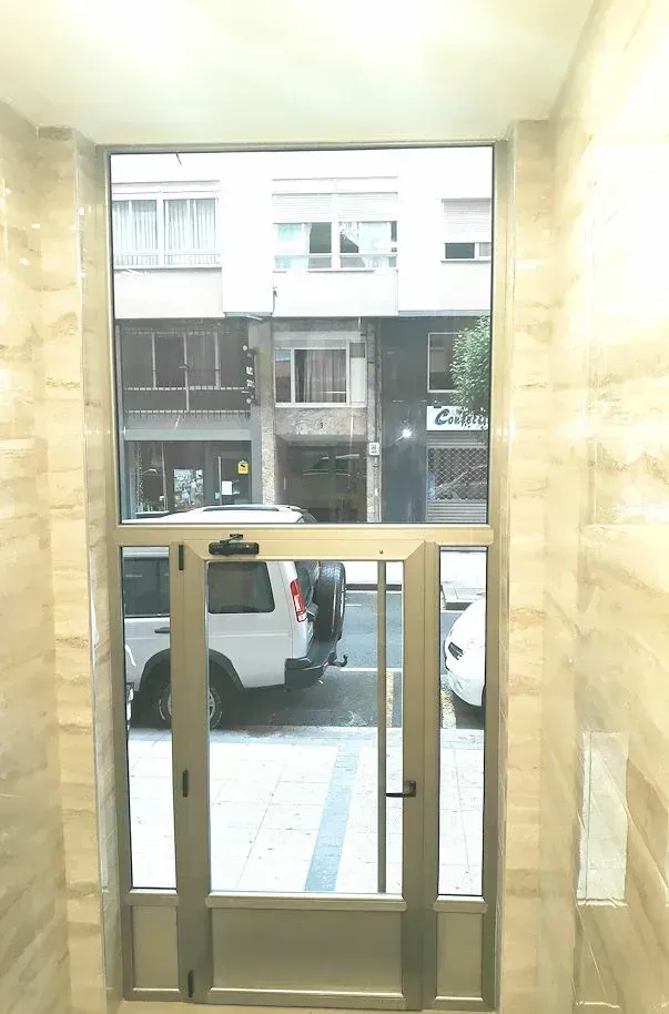 Una puerta y una ventana de cristal dan a una calle de la ciudad. La puerta tiene marco metálico y es automática. Las paredes del portal están alicatadas.