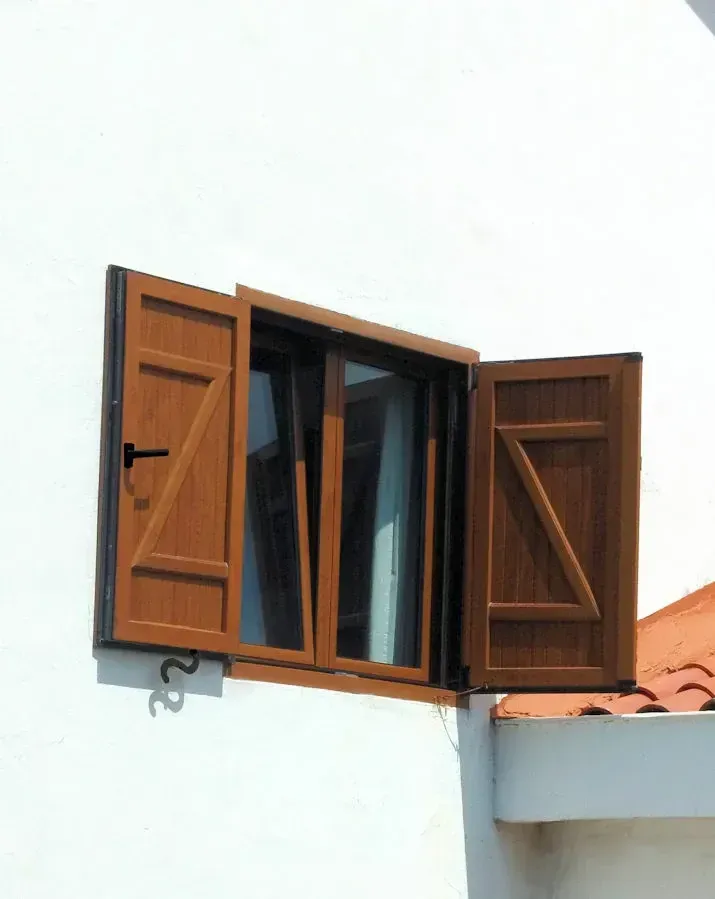 Unas contraventanas marrones se abren en una ventana de un edificio blanco, desde donde se vislumbra un techo de tejas rojas debajo.