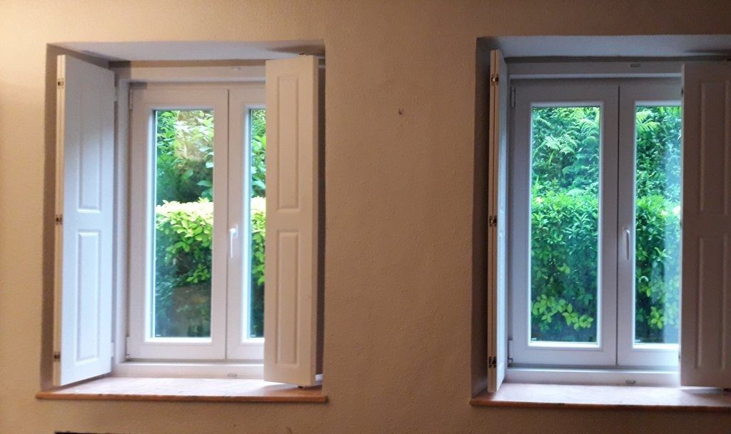Dos ventanas blancas con contraventanas abiertas en una pared de color claro, que enmarcan una vista del follaje verde del exterior.