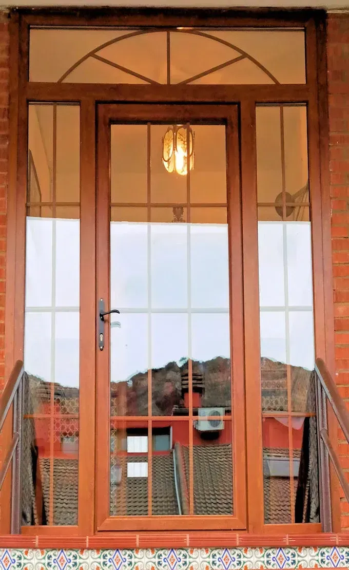 Puerta principal con marco marrón, paneles de vidrio y ventanas laterales que reflejan un edificio de tejado rojo. Una pequeña ventana arqueada se encuentra sobre la puerta.