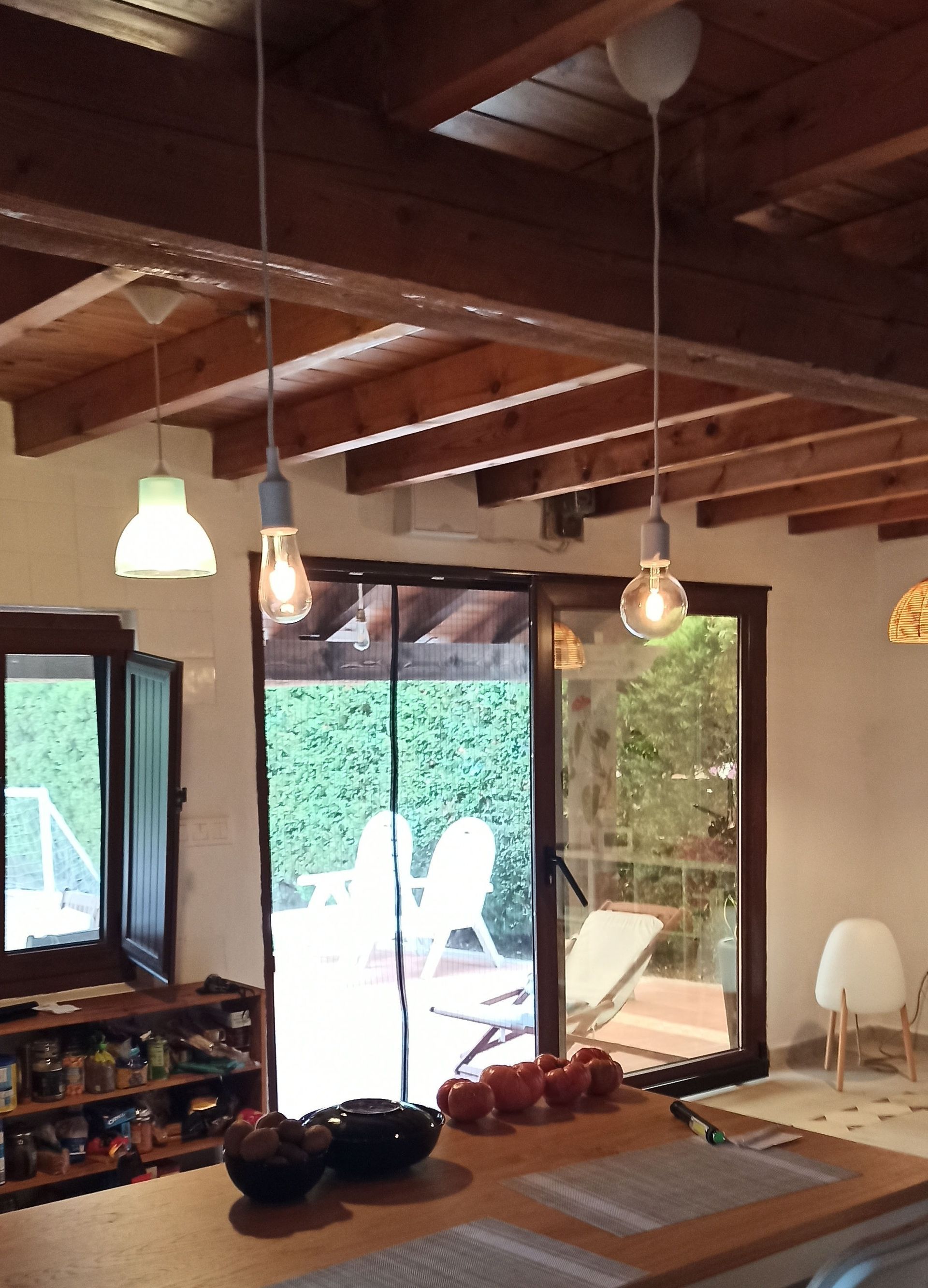 Cocina con vigas de madera, lámparas colgantes y vista al jardín a través de una puerta corrediza de vidrio. Mostrador con fruta en primer plano.