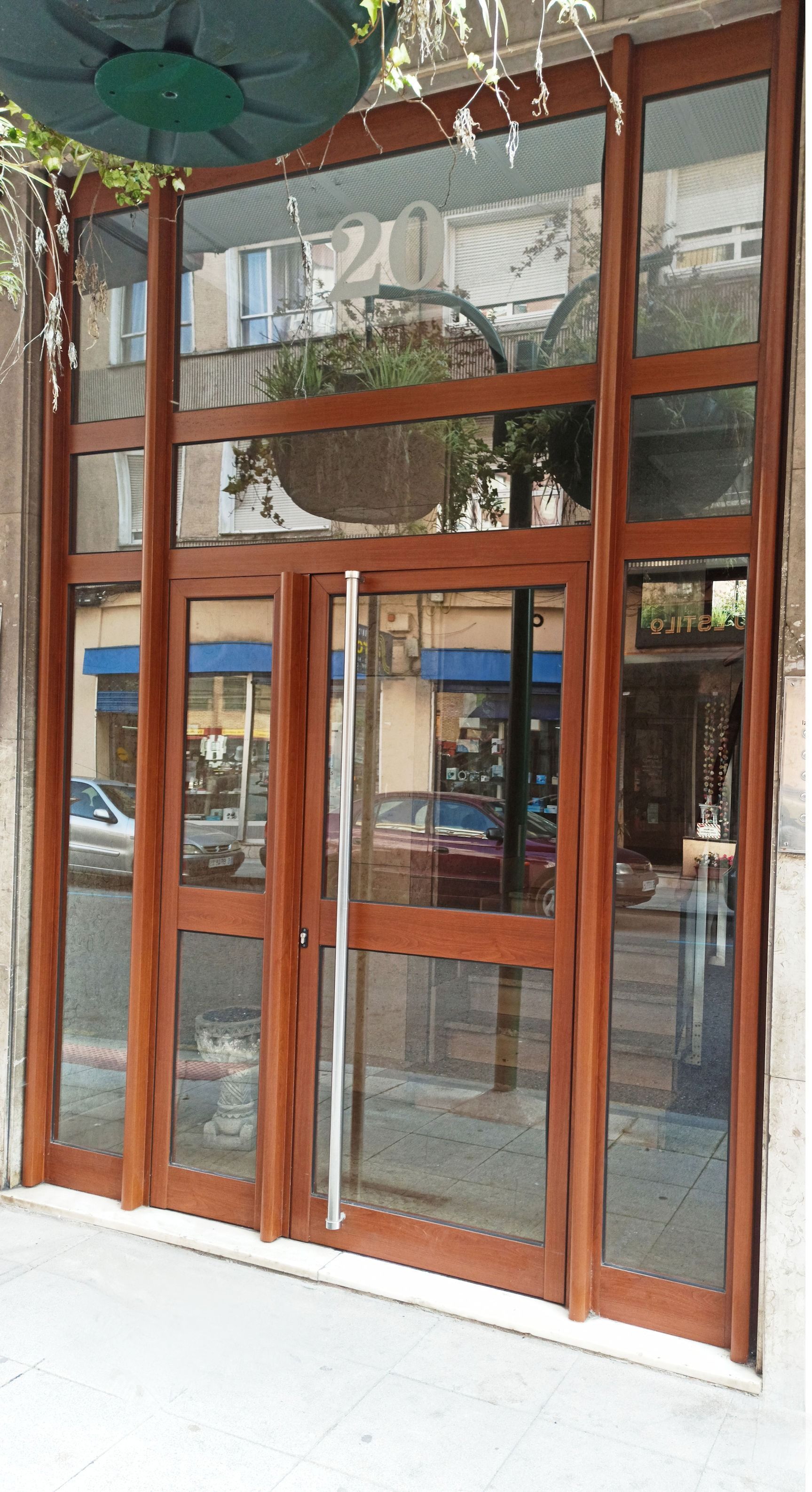 Puertas y ventanas de vidrio con marco de madera de una tienda. Una planta colgante sobre la entrada refleja una escena callejera.