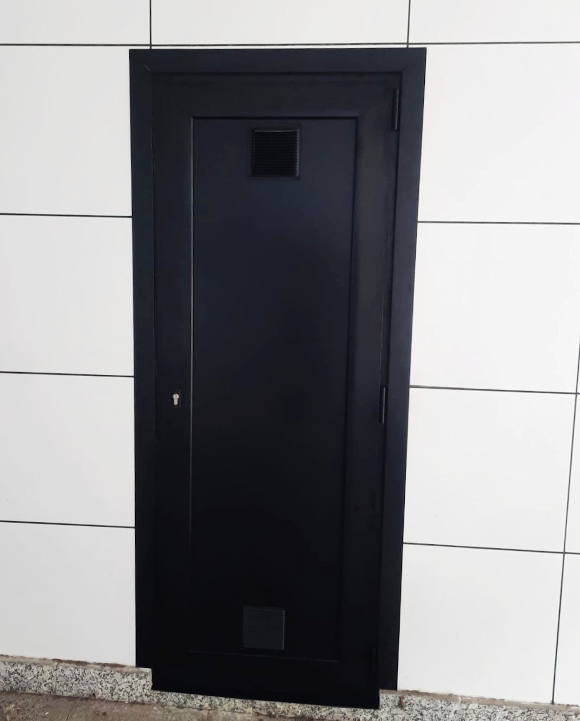 Puerta negra empotrada en una pared de azulejos blancos con una pequeña ventana cuadrada encima y debajo de la cerradura.
