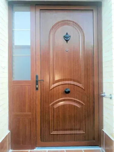 Puerta de entrada de madera con paneles decorativos y ventana lateral. Diseño ornamentado en marrón oscuro con aldaba y tirador.