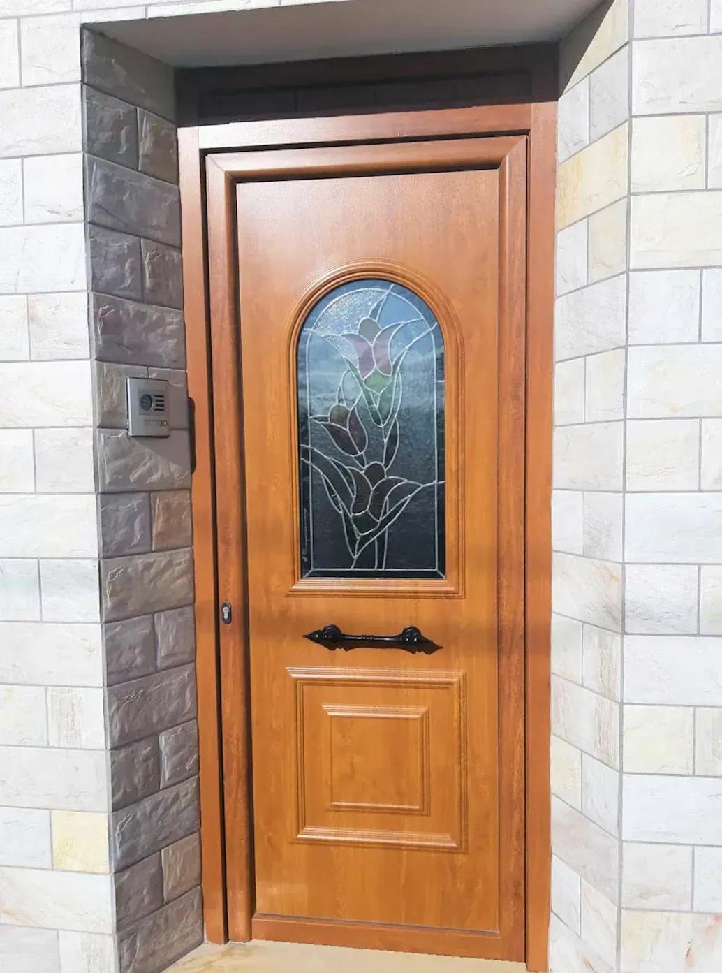 Puerta de madera con panel de vidrio arqueado, manija negra y paneles decorativos, colocada en una entrada de ladrillo.