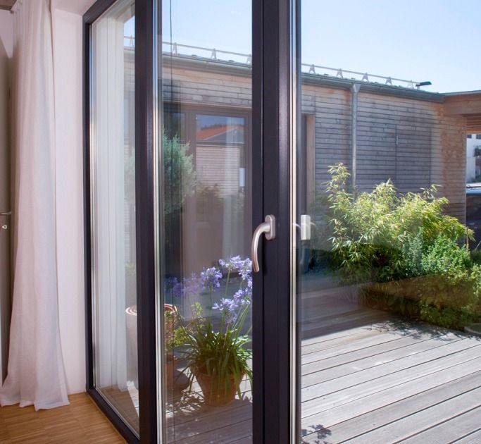 Puerta corrediza de vidrio abierta a una terraza de madera con plantas en macetas y un edificio, interior con cortina blanca.