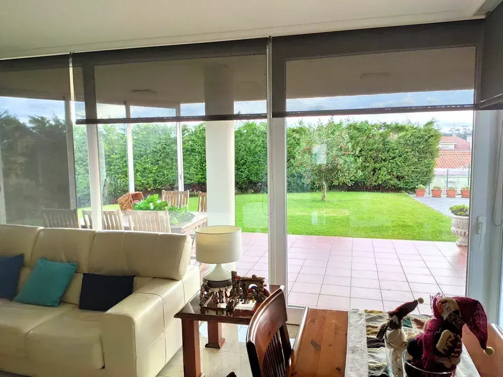 Sala de estar con grandes ventanales, parcialmente cubiertos con persianas oscuras, con vistas a un patio trasero con vegetación. En el interior se ven un sofá beige y una mesa.