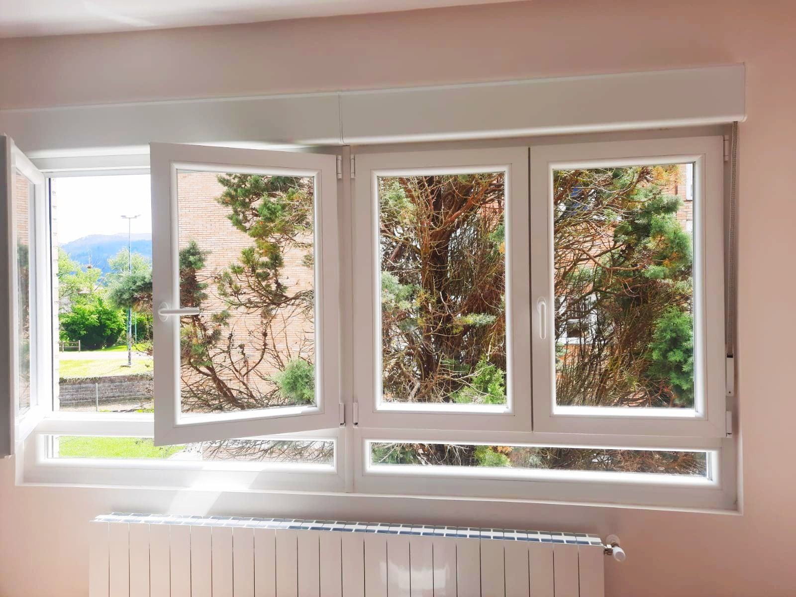 Una ventana blanca de varios paneles, con una sección abierta, que muestra una vibrante vista exterior de árboles y una montaña bajo un cielo claro.