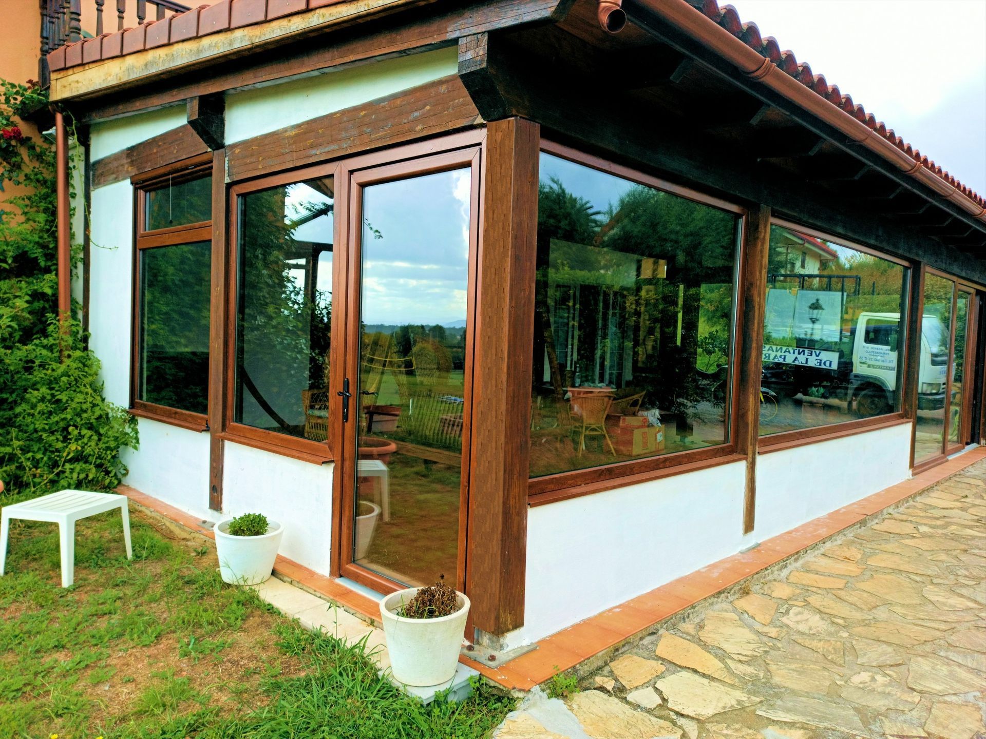 Un solárium con paredes de cristal y marco de madera marrón, rodeado de césped y un patio de piedra. Cerca hay una pequeña mesa blanca y macetas con plantas.
