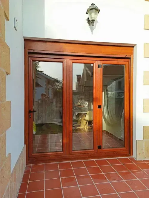 Una puerta de vidrio de tres paneles con marco de madera, que refleja escenas del exterior. La puerta da a un patio de baldosas rojas, bajo una pared blanca con una luz decorativa.