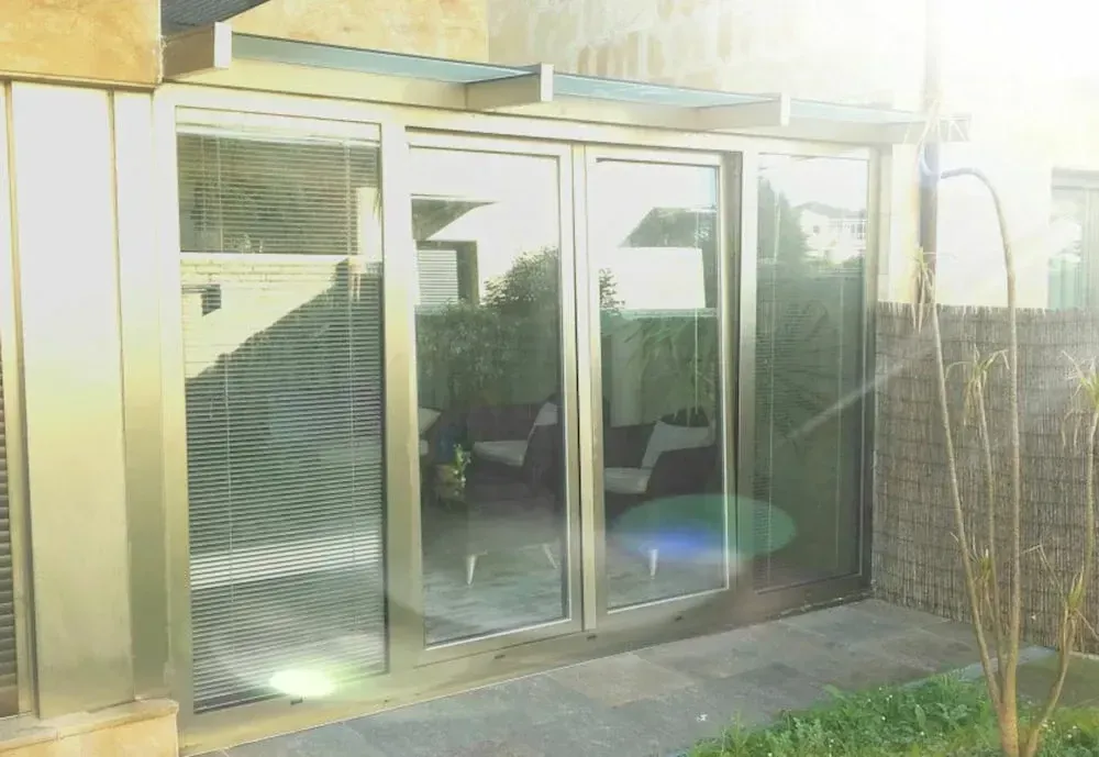 Unas puertas de vidrio con marcos plateados dan acceso a un patio con vegetación y asientos. La luz del sol se refleja en el cristal.
