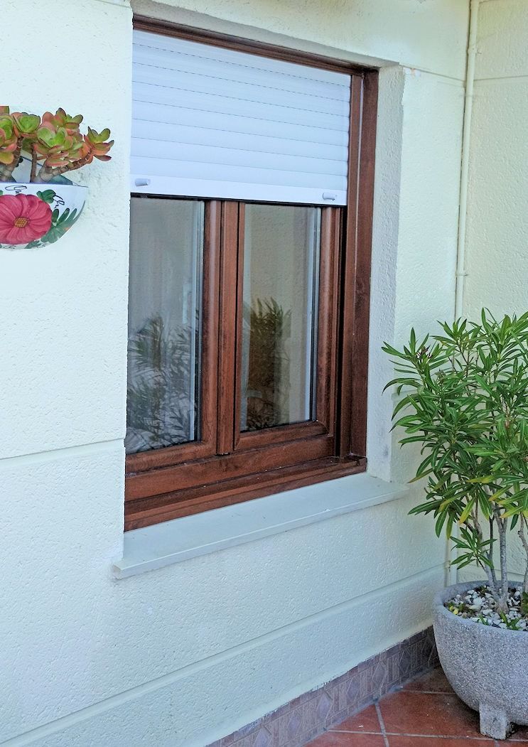 Ventana de marco marrón con persianas blancas, entreabierta, en un edificio color crema. Junto a la ventana hay una planta en maceta.