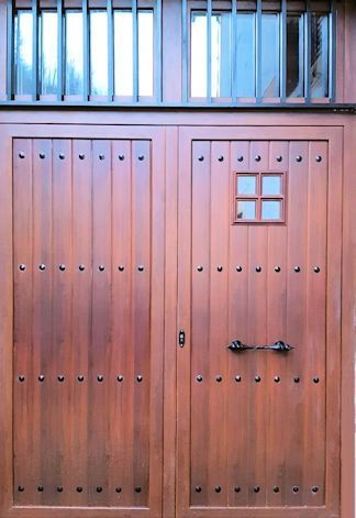 Puertas dobles de madera con detalles tachonados y una pequeña ventana cuadrada; arriba, dos ventanas con rejas negras.