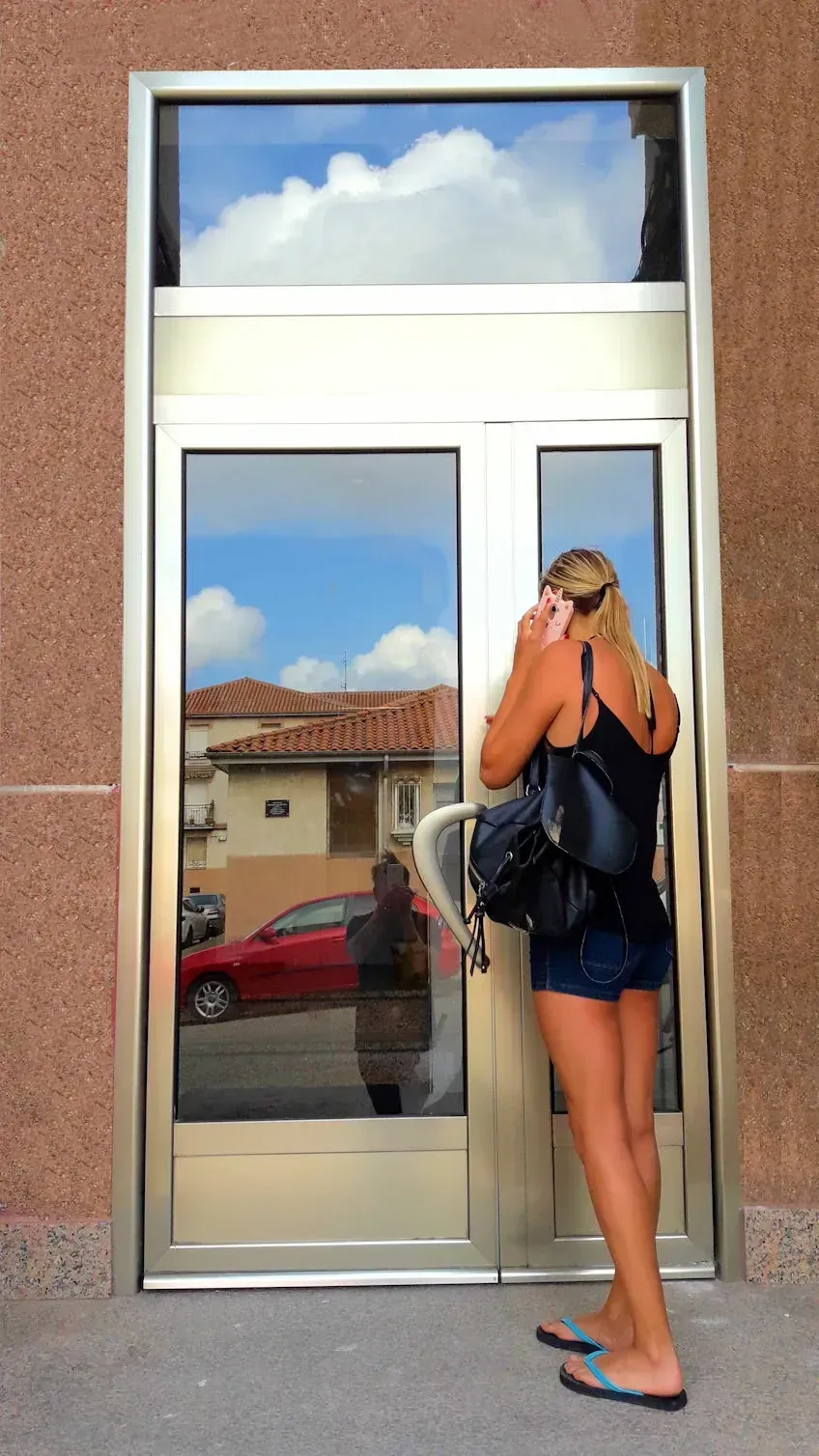 Mujer en pantalones cortos y camiseta sin mangas hablando por teléfono, parada afuera de un edificio con puertas de vidrio, que reflejan el cielo y la calle.