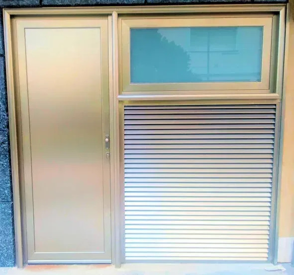 Puerta exterior de color bronce con ventana y persianas horizontales, probablemente una entrada.