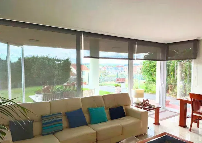 Sala de estar con grandes ventanales, un sofá beige con cojines coloridos, una mesa de comedor y persianas enrollables gris oscuro. Se puede apreciar una vista de la vegetación y la ciudad a lo lejos.