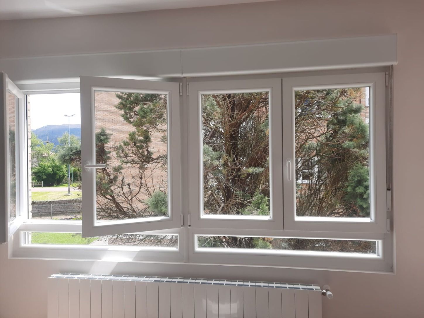 Ventanas con marco blanco, una de ellas abierta, con vistas a la vegetación y a las montañas lejanas. Un radiador se encuentra debajo de las ventanas.
