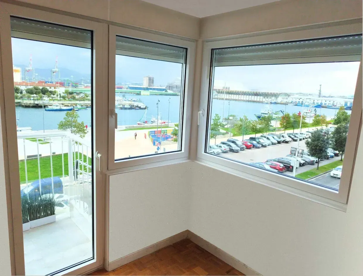 Habitación esquinera con amplios ventanales con vistas a un puerto con barcos, coches y gente. Paredes y molduras blancas, suelo de madera.