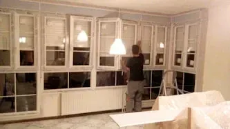 Persona instalando una luminaria cerca de una serie de ventanas con marco blanco y un radiador en una habitación.