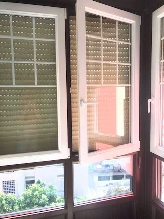 Ventana con marco blanco parcialmente abierta, con vista al exterior. Tiene un patrón de cuadrícula y un parasol.