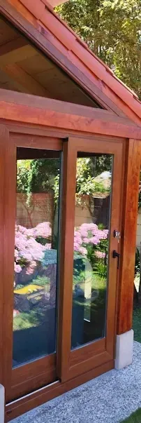 Puertas de vidrio con marco de madera de una pequeña estructura de jardín con techo inclinado. Jardín verde y flores rosas se reflejan en el cristal.
