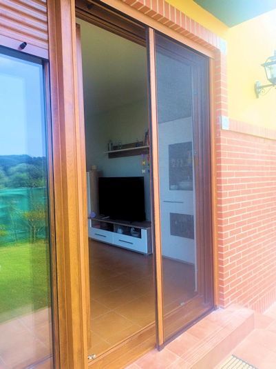 Puerta corrediza de vidrio con mosquitera retráctil que da acceso a la sala de estar. El marco de la puerta es marrón, con una vista de un televisor y muebles blancos.